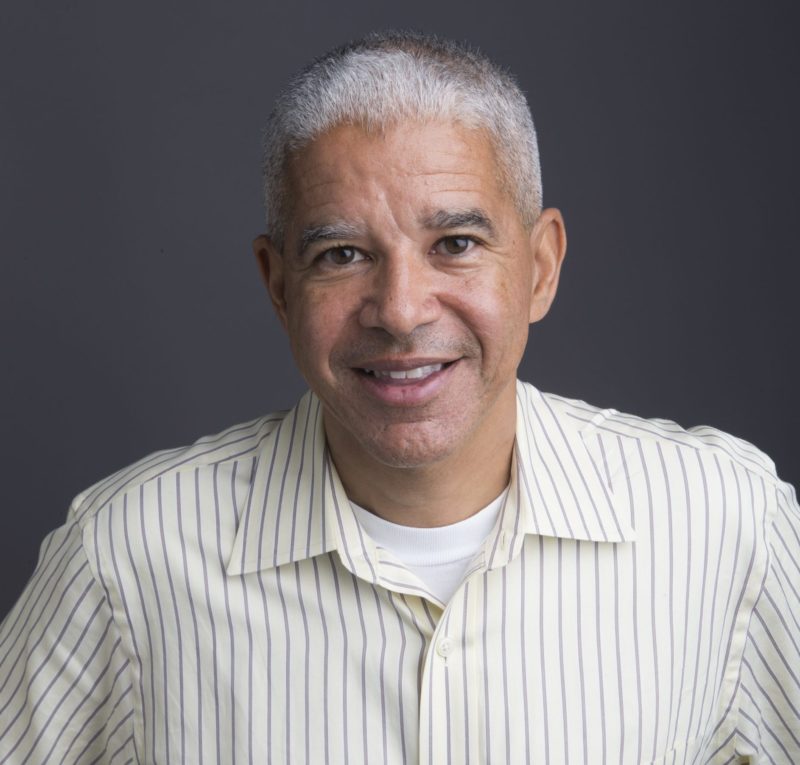 A middle-aged man with short gray hair smiles while looking at the camera. He is wearing a light-colored, striped dress shirt against a plain dark background, embodying the thoughtful demeanor reflective of Berkeley Journalism's seasoned professionals.