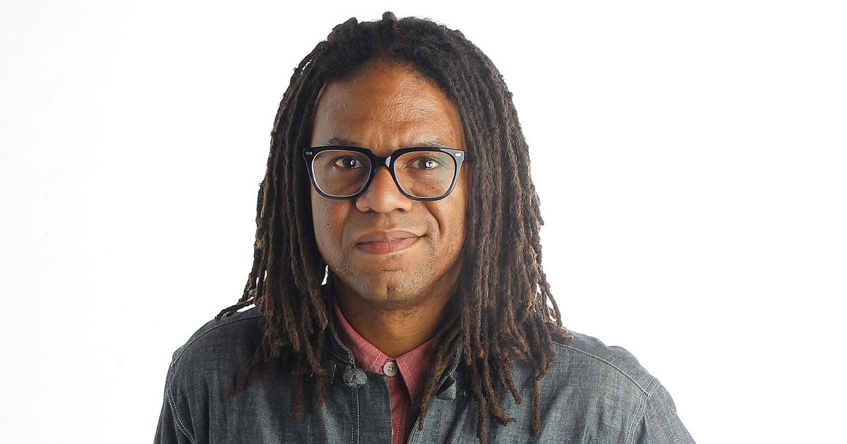 A man with shoulder-length dreadlocks and black-rimmed glasses, embodying the Berkeley Journalism vibe, is wearing a denim shirt over a red collared shirt. He has a neutral expression and is standing against a plain white background.