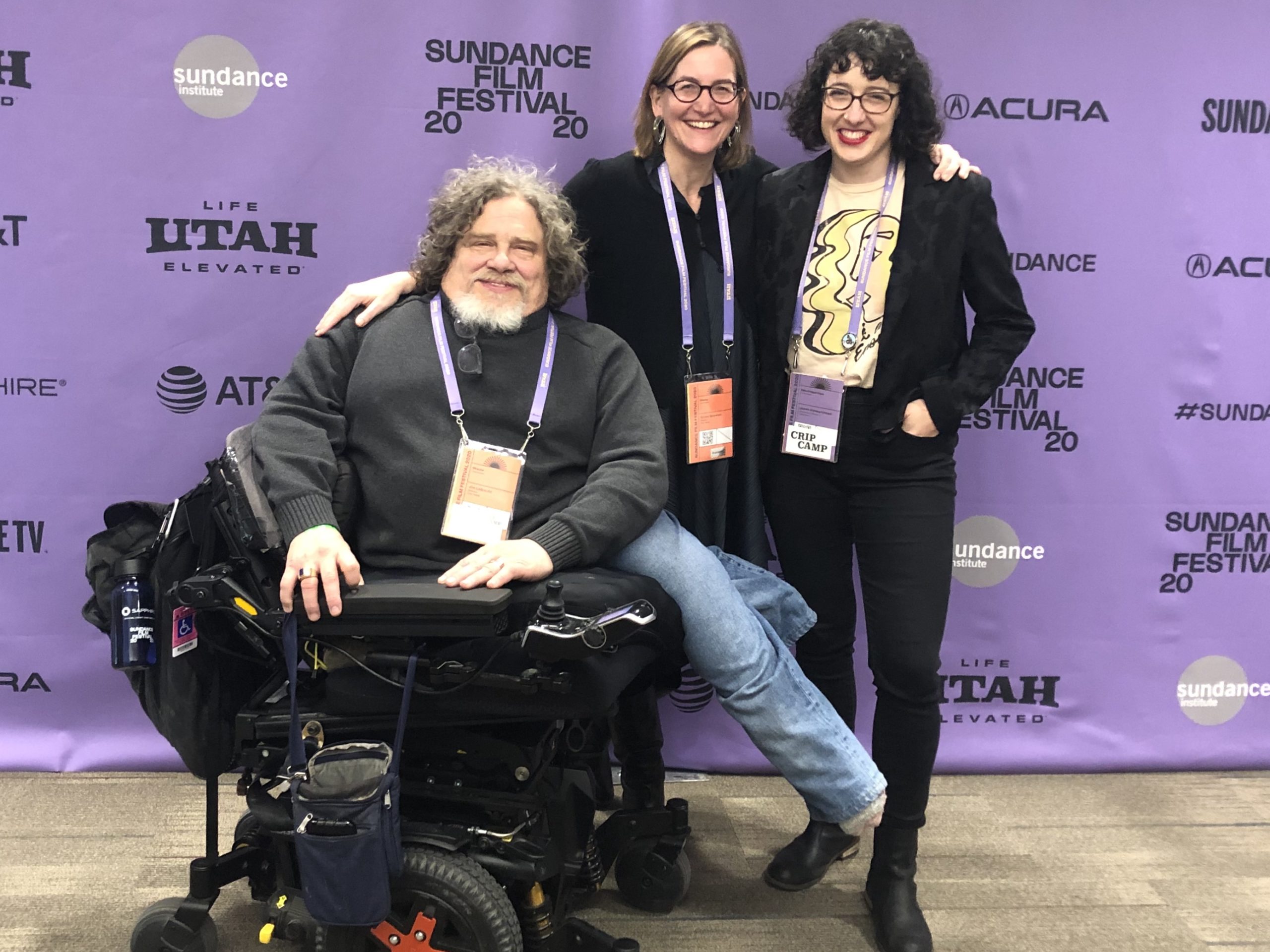 Three people pose together at the Sundance Film Festival against a purple backdrop with logos. The person on the left sits in a wheelchair, while the two others stand beside them, smiling. All wear festival badges and look cheerful, embodying the spirit of Berkeley Journalism's diverse storytelling.
