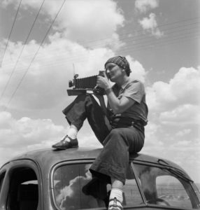 A woman sits on the roof of a vintage car, aiming a large format camera. She is wearing a headscarf, short-sleeve shirt, high-waisted pants, and socks with sandals. The sky above her is filled with scattered clouds and power lines in the background—an intriguing scene straight out of a Berkeley Journalism project.