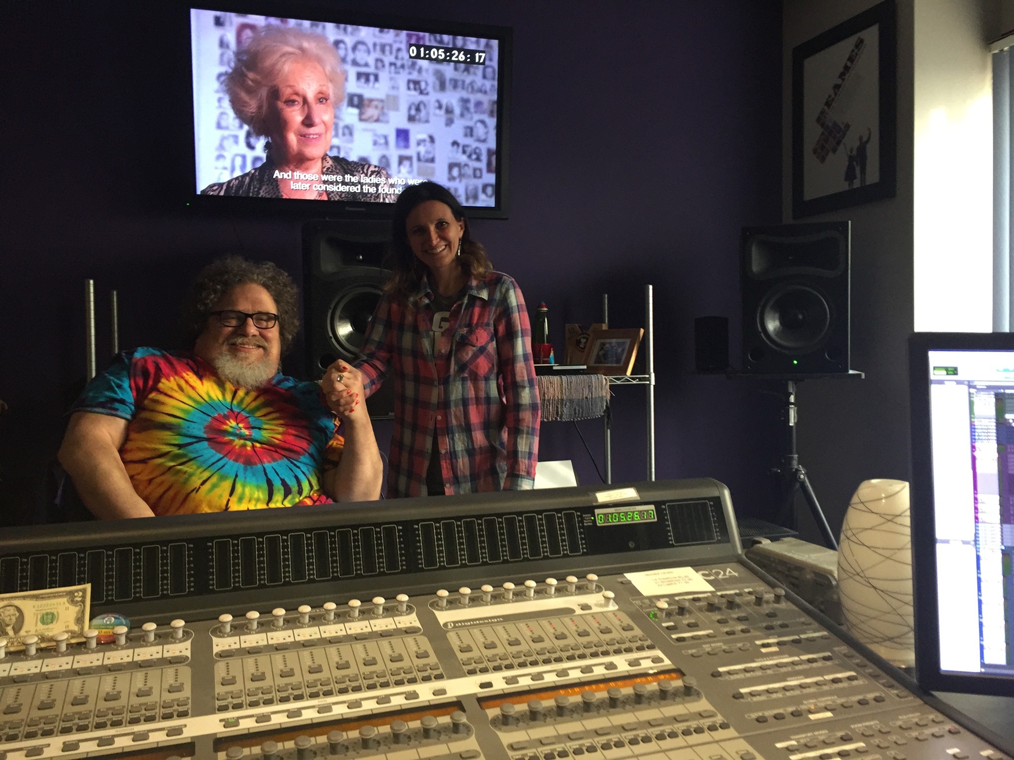 Two people are in a recording studio at Berkeley Journalism. One person, wearing a colorful tie-dye shirt, is seated, while the other, in a plaid shirt, stands beside them. A sound mixing console dominates the foreground, and a television screen shows an elderly woman in the background.