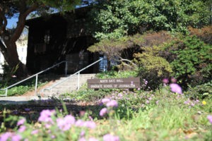 A pathway bordered by greenery and flowers leads to North Gate Hall, home to Berkeley Journalism's Graduate School of Journalism. A sign with these details is visible amidst the plants, and the hall is partially obscured by shadows cast by trees and sunlight.