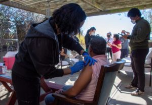 A healthcare worker in gloves and a mask administers a vaccine to a seated individual under a shaded outdoor area, an effort captured vividly by Berkeley Journalism students documenting the scene. Nearby, several masked people stand and sit, engaging in various activities.