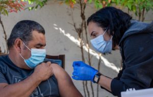 A man sits outdoors, pulling up his sleeve as a healthcare worker wearing a mask and blue gloves administers a vaccination shot into his upper arm. Both individuals are focused on the procedure, with green plants in the background. Documenting this moment is Berkeley Journalism capturing the essence of public health efforts.