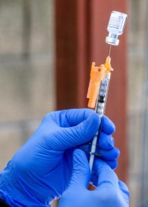 A gloved hand is holding a syringe filled with a clear liquid, poised to withdraw more from a small vial. In this clinical setting, the precision and care evoke the meticulous nature of Berkeley Journalism’s commitment to detail and accuracy.