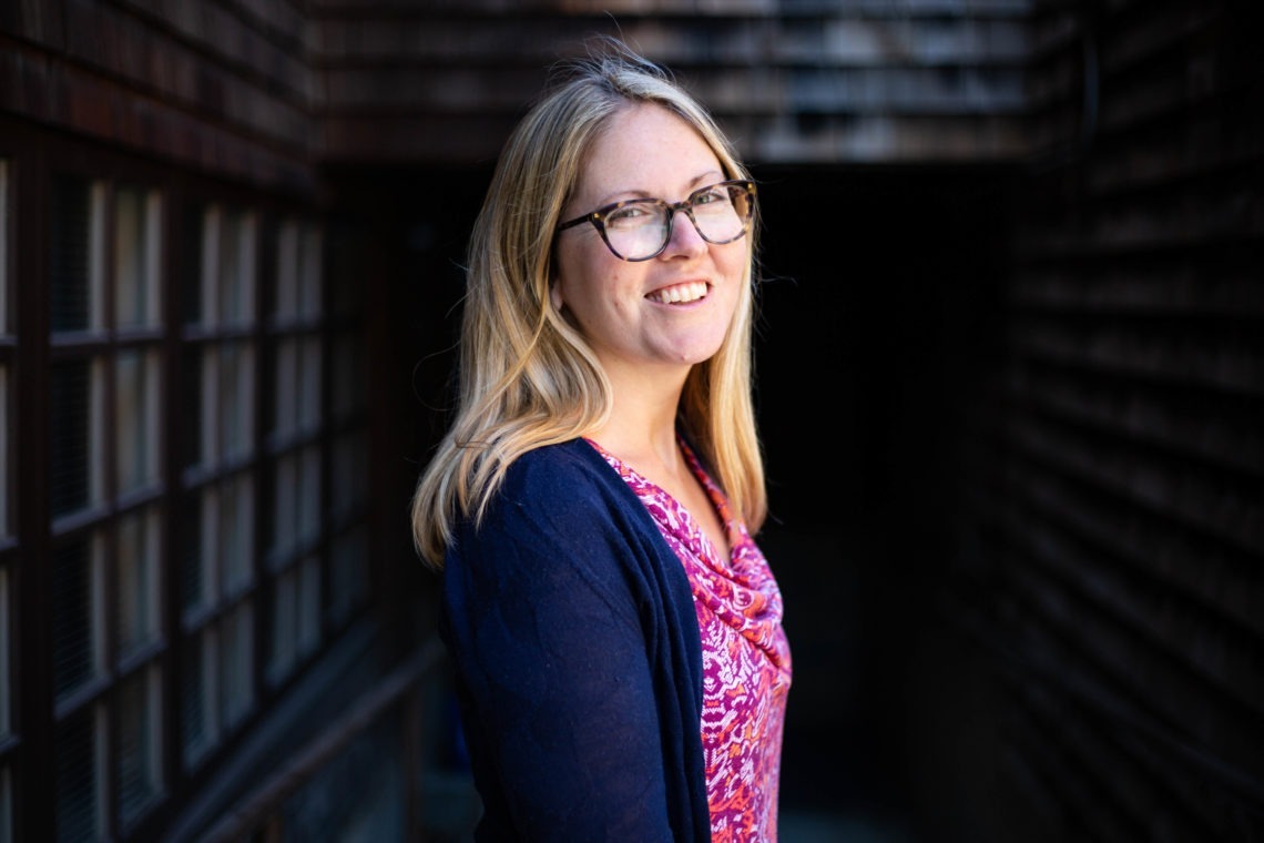 A woman with long blonde hair and glasses stands outdoors in a narrow pathway between wooden buildings. She is smiling and wearing a dark cardigan over a pink patterned blouse, embodying the energetic spirit of Berkeley Journalism.