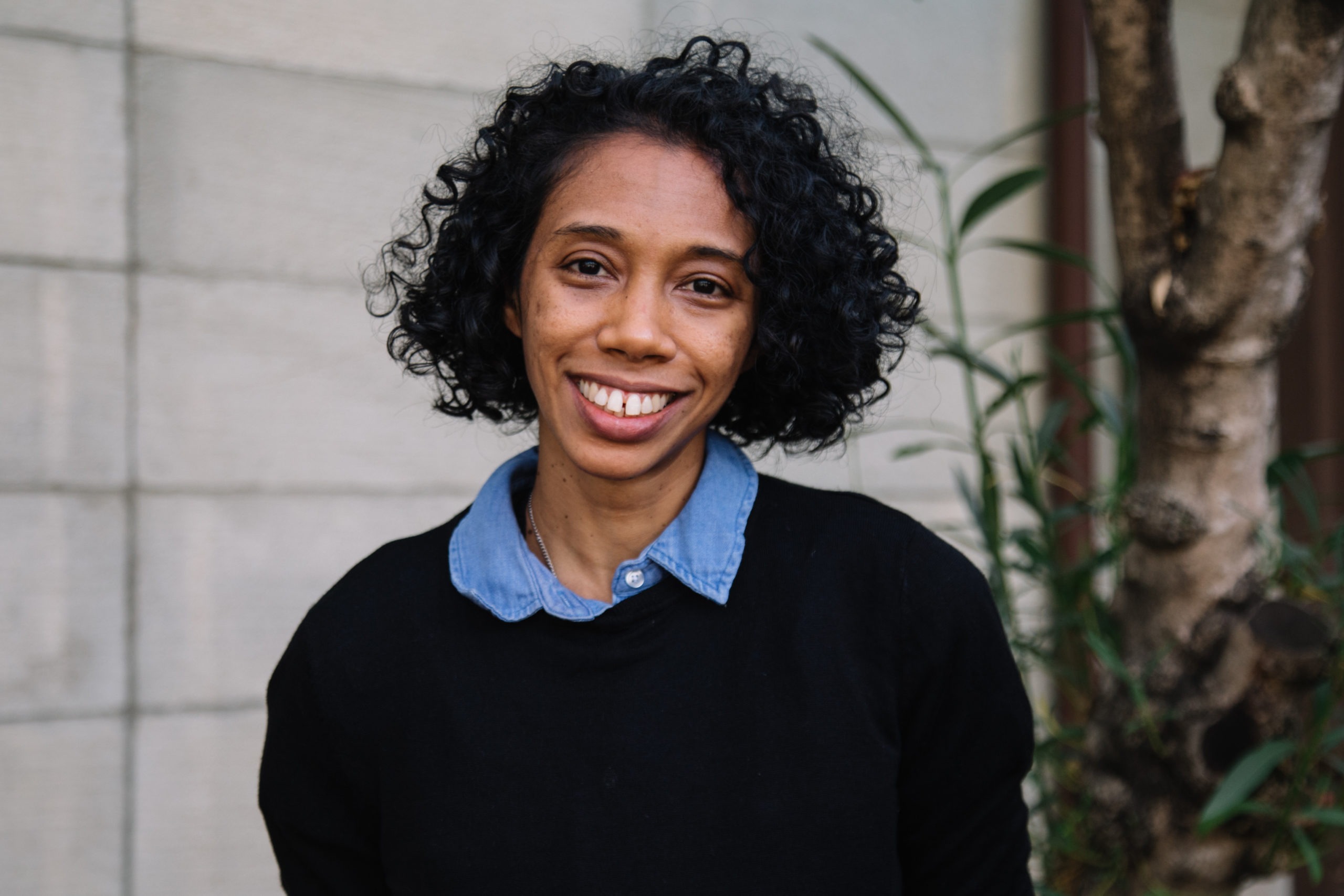 A person with short curly hair smiles warmly at the camera. They are wearing a black sweater over a blue collared shirt. The background, reminiscent of Berkeley Journalism's dynamic environment, features a gray brick wall and some greenery.