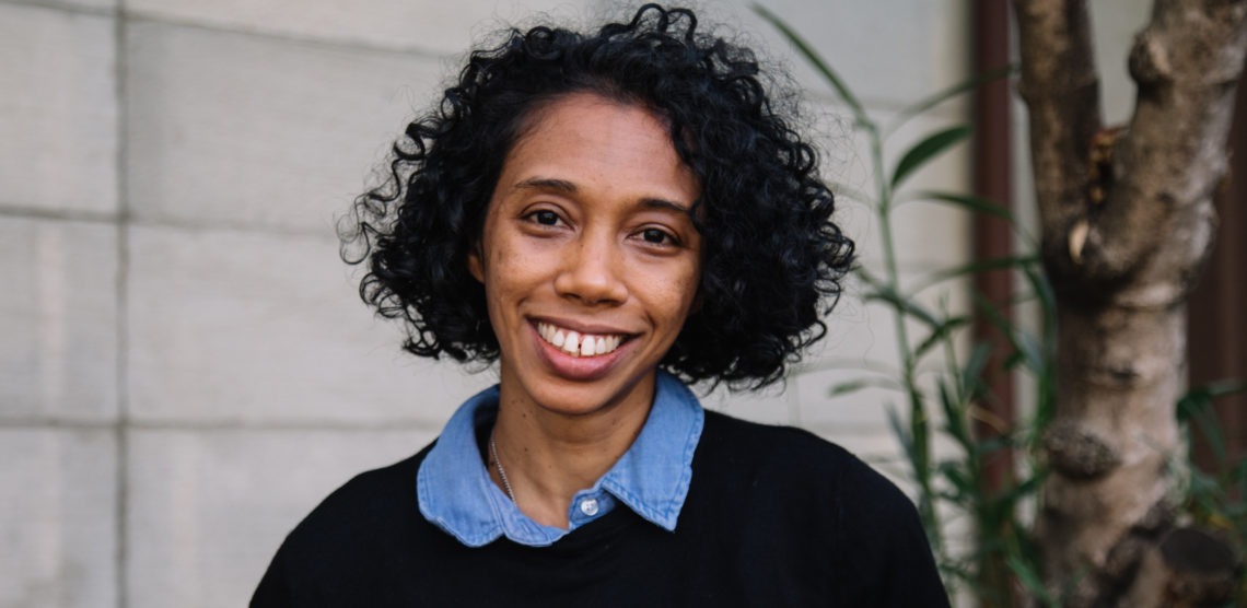 A smiling person with curly dark hair standing outdoors. They are wearing a blue collared shirt under a black sweater. A tree and part of a building wall from the Berkeley Journalism campus are visible in the background.
