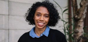 A smiling person with curly dark hair standing outdoors. They are wearing a blue collared shirt under a black sweater. A tree and part of a building wall from the Berkeley Journalism campus are visible in the background.