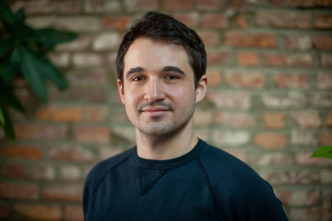 A man with short dark hair and a light beard, possibly a Berkeley Journalism student, smiles softly while standing in front of a brick wall. He is wearing a dark, long-sleeved shirt. There is some greenery visible on the left side of the image.