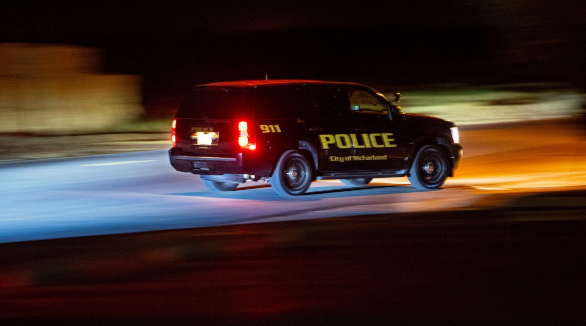 Police SUV with "City of McFarland" text and 911 emblem driving quickly on a road at night, its lights casting blue and amber glows. With the background blurred due to the vehicle