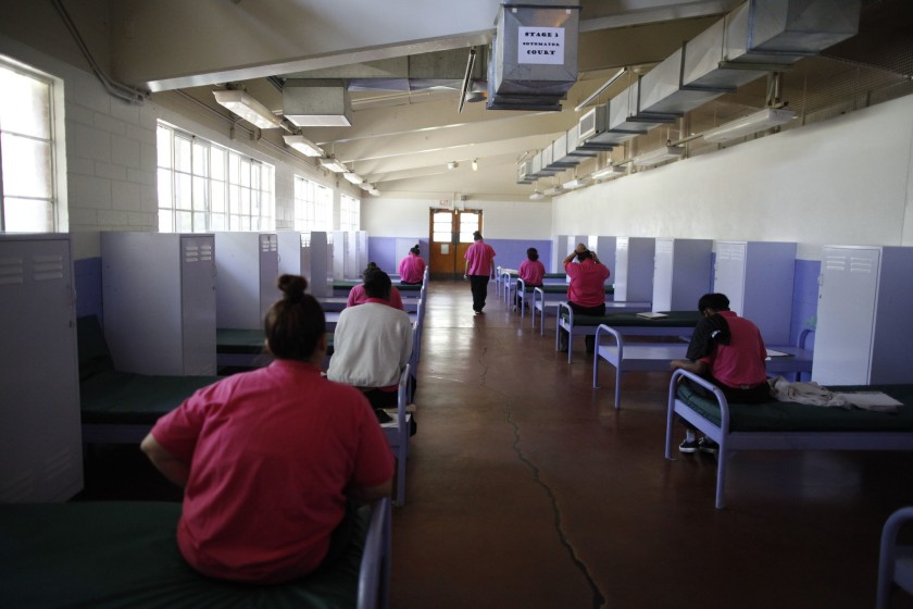 A large room with multiple beds, each separated by partitions. Several individuals sit on the beds, wearing pink uniforms. The room has white walls, with a ceiling featuring exposed ductwork and fluorescent lighting. A sign on the ceiling reads, "COMMUNITY CENTER," bringing to mind snapshots from a Berkeley Journalism feature.