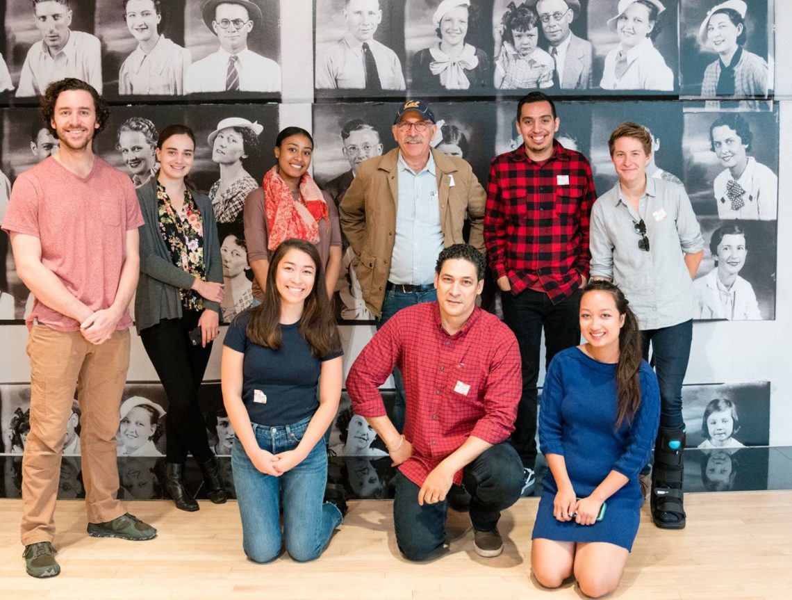 A group of ten people posing and smiling in front of a collage of black and white photos on a wall. They are casually dressed, with some kneeling in front while others stand behind. The historical portraits on the background wall emphasize the rich heritage associated with Berkeley Journalism.