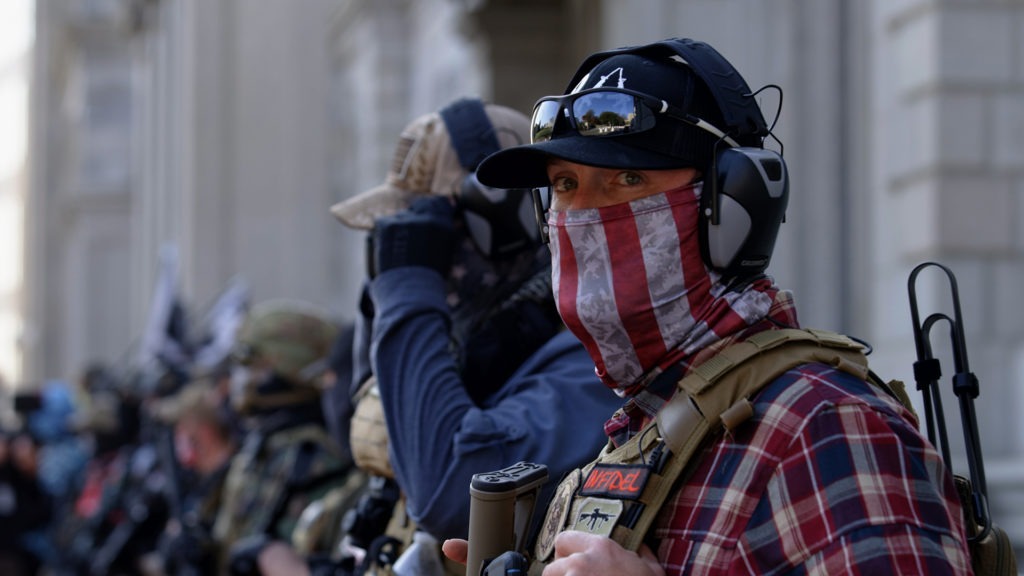 A group of people, some in tactical gear and masks, standing outside a building. One person in the foreground is wearing a red, white, and blue face covering, sunglasses, and a cap with an earpiece and communication device visible. They appear to be participating in a demonstration or gathering covered by Berkeley Journalism.