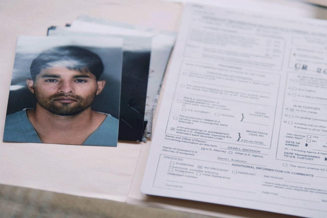 A photograph of a man with dark hair and a beard is placed next to several documents on a table, resembling forms often used in Berkeley Journalism. The documents appear to be legal or identification forms with various fields and checkboxes filled in.