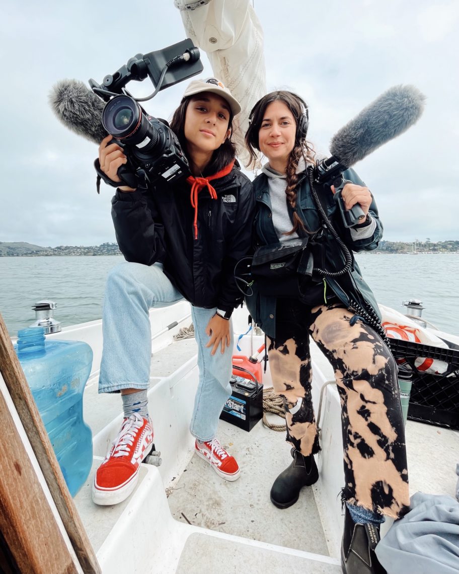 Two young adult woman standing in a stylish pose while holding camera and sound equipment. They are on a small boat in the ocean. 