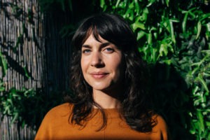 A woman with shoulder-length brown hair and bangs smiles slightly, standing outdoors in front of a lush green foliage background. Clad in an orange sweater, she squints slightly in the sunlight. Her demeanor embodies the thoughtful curiosity often seen in Berkeley Journalism students.