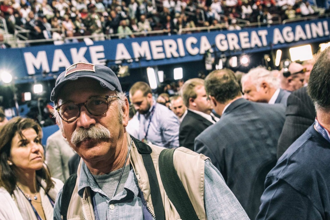 A man with a mustache wearing a cap and glasses stands in the foreground of a crowded event. Behind him, there is a large banner with the slogan "Make America Great Again!" Amidst the busy atmosphere, reminiscent of Berkeley Journalism gatherings, a diverse group engages in lively conversations.