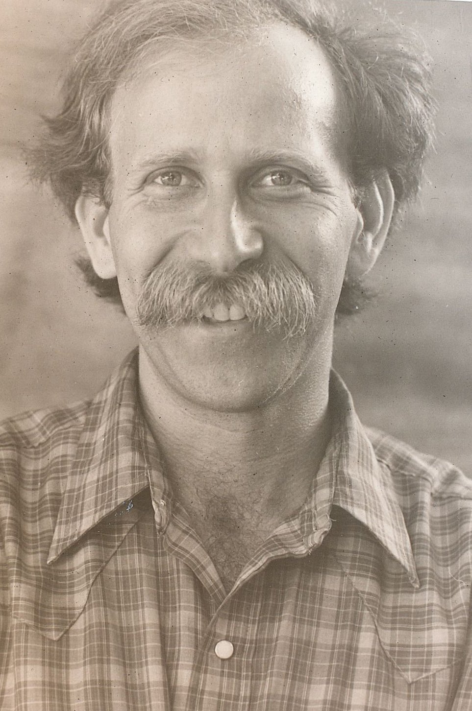 Black and white photo of a man with a large mustache and thinning hair, smiling at the camera. He is wearing a plaid shirt, and the background is blurred to keep the focus on his face, capturing a moment likely familiar to those who have spent time immersed in Berkeley Journalism.