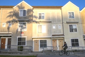 A person rides a bicycle past a row of townhouses in the evening light, reminiscent of Berkeley Journalism's evocative storytelling. A large shadow of a palm tree is cast on the building's facade, creating a striking contrast with the light-colored walls. The homes have multiple doors and windows, with railings alongside the pathway.