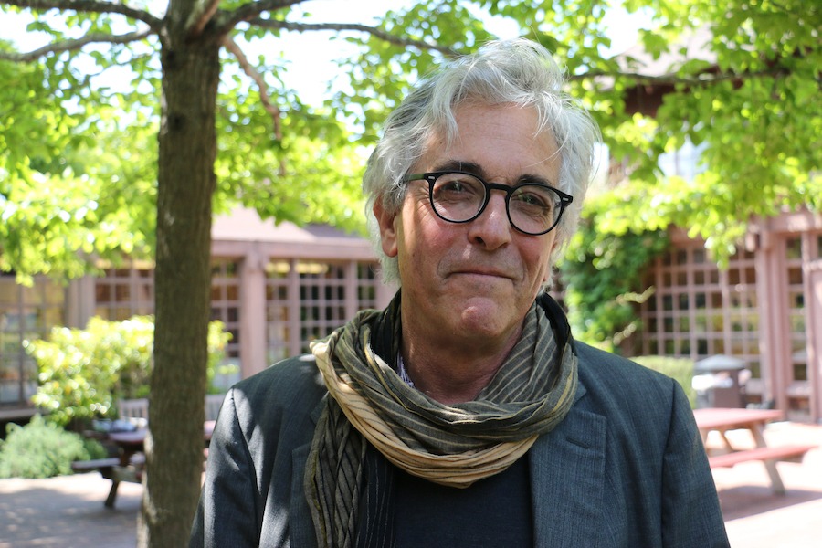 Photo of a man with grey cool hair wearing black glasses, a scarf and a suit jacket in the courtyard of North Gate Hall.