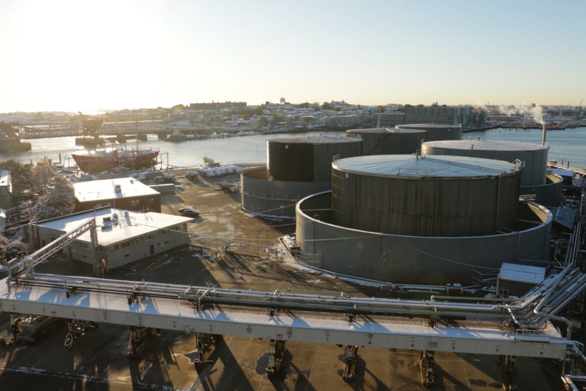 Aerial view of an industrial waterfront facility. Large cylindrical storage tanks dominate the foreground, with buildings and pipes surrounding them. In the background, a harbor with anchored ships and city buildings bathed in early morning sunlight is visible, reminiscent of Berkeley Journalism's insightful coverage.
