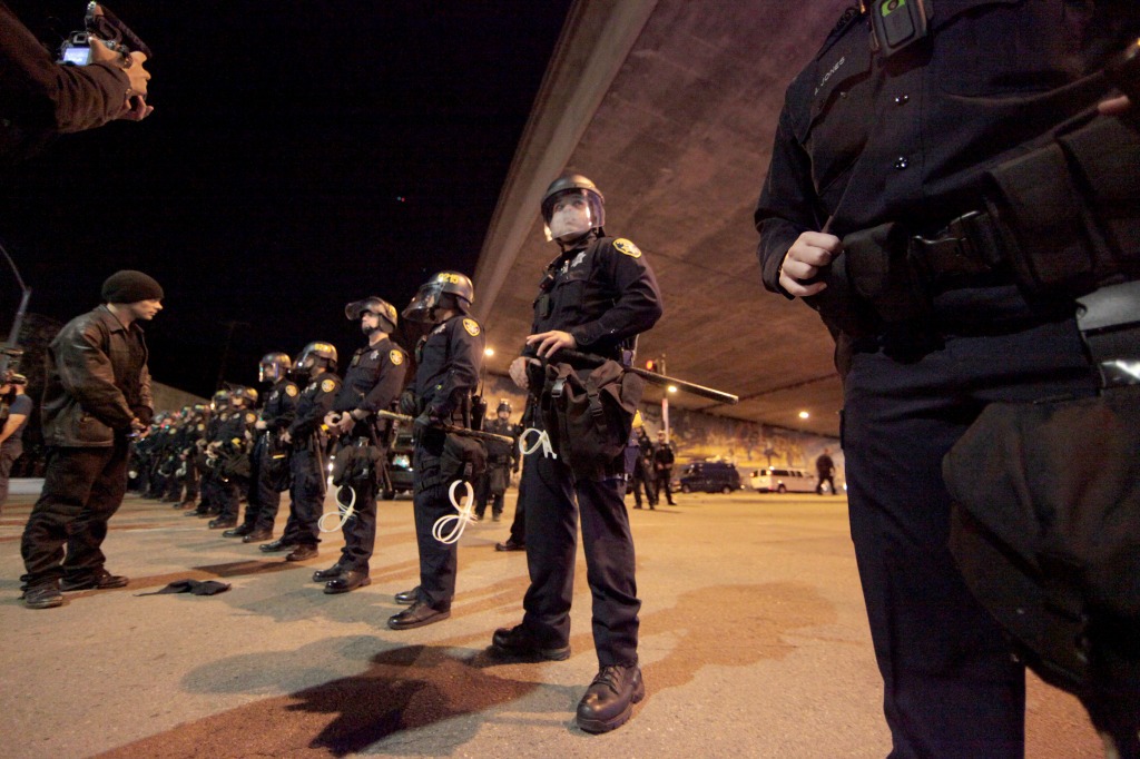 Police officers in riot gear stand in a line under a highway overpass at night, an image captured by Berkeley Journalism. Some officers are holding batons and zip tie handcuffs. A few individuals are visible on the left, facing the officers. The scene appears tense yet organized.