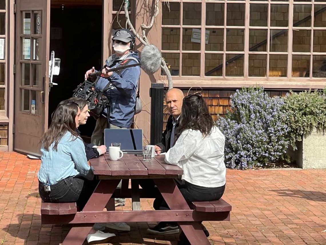 Three people are seated at a wooden picnic table outside, engaged in conversation with a laptop open in front of them. A person with a camera and boom microphone stands nearby recording the scene for a Berkeley Journalism project. The background features a large window and a brick pathway.