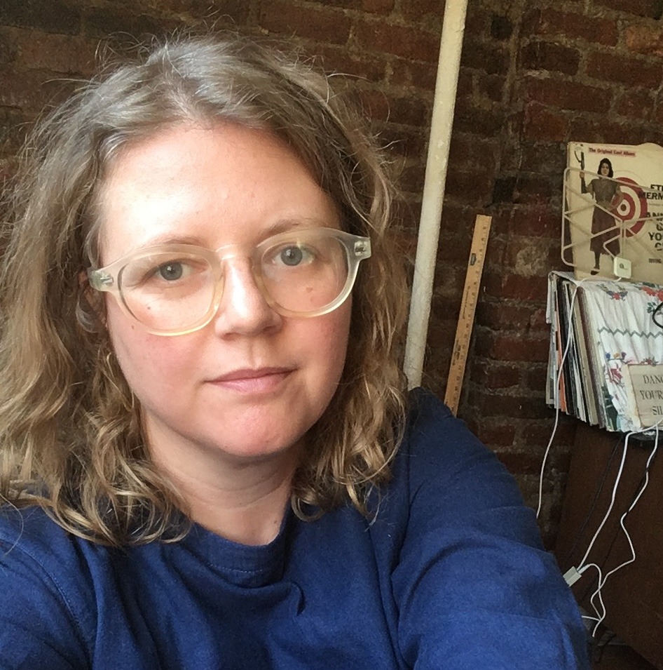 A person with shoulder-length wavy hair and glasses is wearing a blue shirt. They are indoors with a brick wall, a magazine rack, and several cables visible in the background—reminiscent of a Berkeley Journalism setting. The person is looking directly at the camera with a neutral expression.