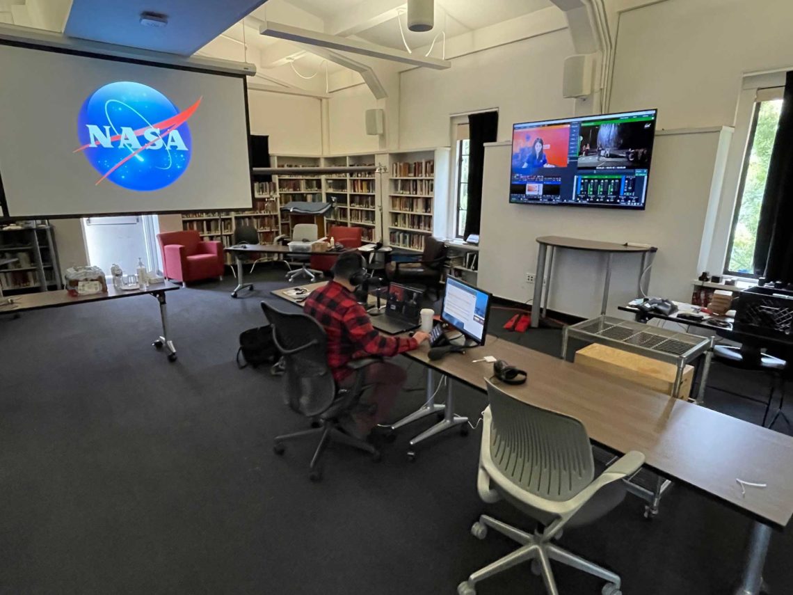 A person sits at a desk in a room with multiple computer monitors and AV equipment. The NASA logo is displayed on a large projector screen. The room, which could be part of a Berkeley Journalism setup, has shelves filled with books and several chairs, creating a modern, tech-equipped workspace or classroom environment.