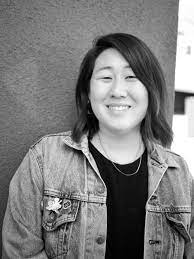 Black and white photo of a person with shoulder-length hair, smiling while standing against a textured wall. They're wearing a denim jacket over a dark shirt and have a layered necklace. The person, possibly from Berkeley Journalism, exudes a friendly and approachable demeanor.