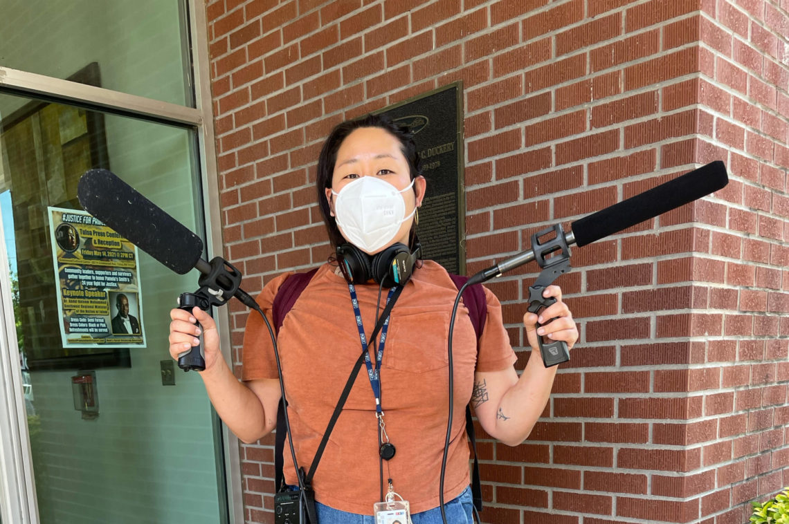 A person wearing a mask holds two microphones outside a brick building. They have headphones around their neck and a lanyard with an ID badge that reads "Berkeley Journalism." A poster and a plaque are visible on the building behind them.