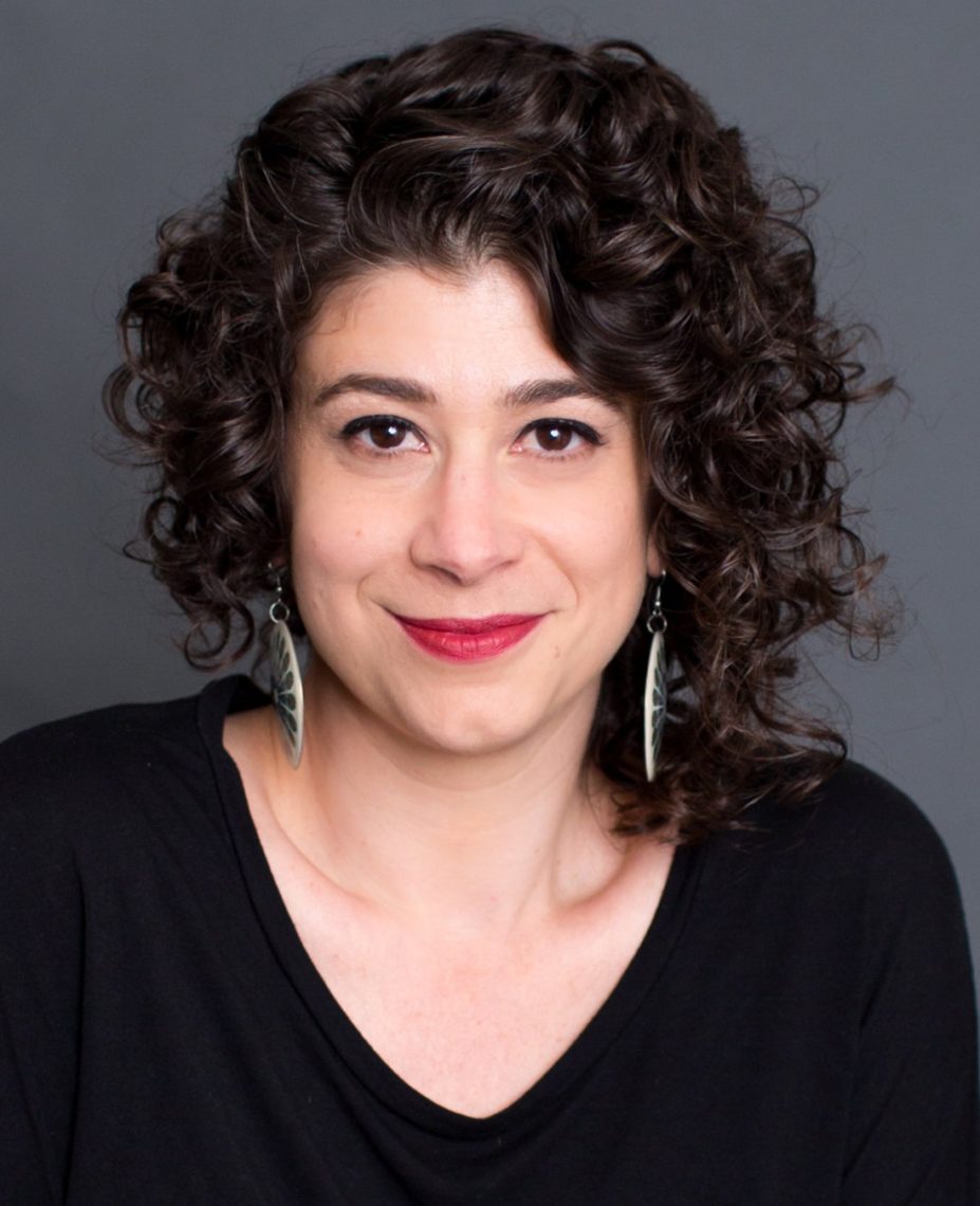 A woman with short, curly dark hair and red lipstick is smiling slightly at the camera. She is wearing a black top and leaf-shaped earrings, embodying the distinct style of Berkeley Journalism. The background is a plain, dark gray color.