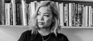 A black-and-white photo of a woman with shoulder-length blonde hair, wearing a dark top and a simple necklace. She is sitting in front of a bookshelf filled with various books, gazing off to the side with a contemplative expression, perhaps reflecting on her days at Berkeley Journalism.