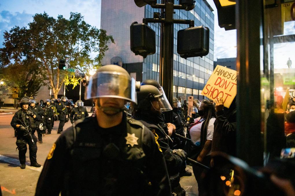 A group of police officers in riot gear stand on a city street, with a crowd of protesters holding signs in the background. One prominent sign reads "HANDS UP, DON'T SHOOT." The scene takes place in front of a modern office building during dusk, highlighted by Berkeley Journalism coverage.