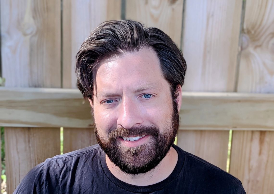 A close up headshot of a middle aged man with medium length hair and a beard. He is sitting in front of a wooden fence.