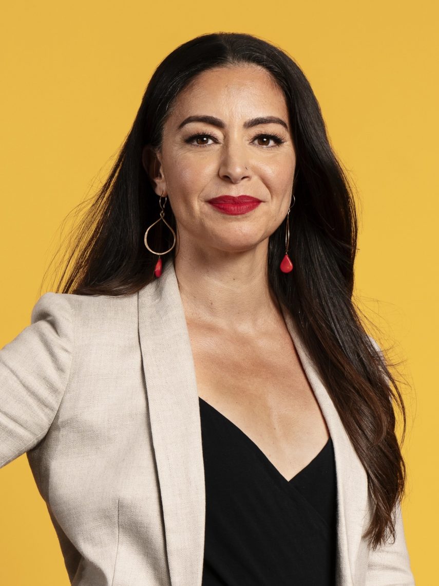A woman with long dark hair stands in front of a yellow background. She is wearing a beige blazer over a black top, red lipstick, and large hoop earrings, one with a teardrop accent. Her expression is confident and she is looking directly at the camera, embodying the bold spirit of Berkeley Journalism.