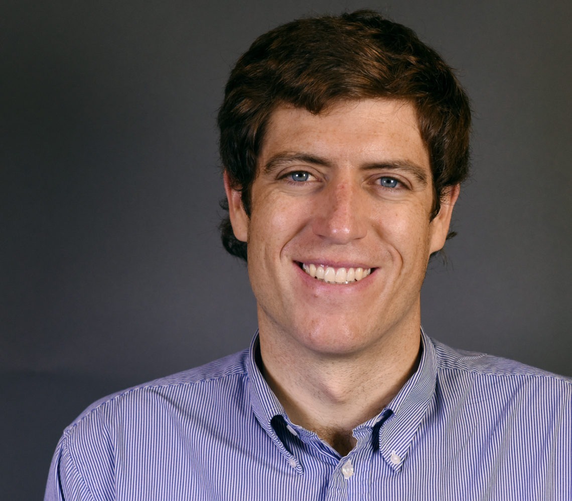A man with short brown hair and blue eyes is smiling at the camera. He is wearing a blue and white striped button-up shirt. The plain, dark background emphasizes his face, giving him a professional aura suitable for Berkeley Journalism.