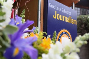 A close-up photo shows a podium with a sign that reads "Graduate School of Journalism" and the emblem of "University of California, Berkeley." Vibrant flowers, including purple and orange blossoms, enhance the foreground, highlighting the prestigious Berkeley Journalism program.