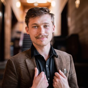 A young man with a well-groomed mustache is confidently smiling at the camera. He is wearing a light brown blazer over a black shirt, standing in a warmly lit hallway with wooden paneling and blurred background, reminiscent of the classic Beryl Terry style.