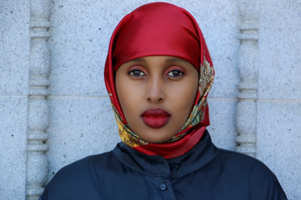 A person wearing a red hijab adorned with a patterned design, and a dark-colored outfit, stands against a textured stone wall. With a serious expression and eyes locked onto the camera, they embody the depth and dedication seen in Berkeley Journalism.