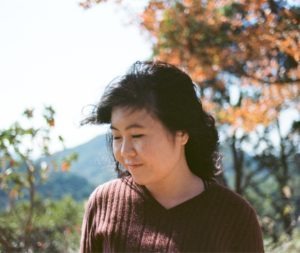 A young woman with dark, wavy hair stands outdoors, looking down and smiling softly. She is wearing a maroon sweater. The background features a blurred mix of green foliage and autumn leaves on trees, with mountains visible in the distance—a perfect scene for a Berkeley Journalism photo essay.