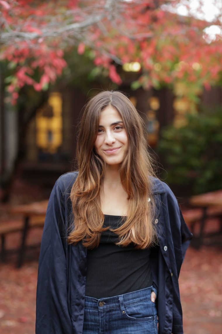  A young woman with long brown hair is pictured wearing a navy blue button up with jeans outside in Berkeley Journalism's courtyard during fall. 
