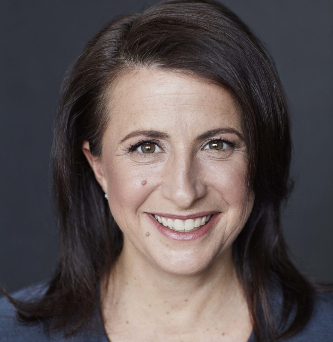A person with shoulder-length dark hair, possibly a Berkeley Journalism student, smiles warmly at the camera. The individual has a light complexion, dark eyes, and is wearing a dark top. The background is plain and dark, putting the focus on the person's face.