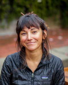 A woman with medium-length dark hair and bangs, wearing a black Patagonia jacket, smiles at the camera. She has a nose ring and a relaxed expression. Behind her, the slightly blurred background features greenery and brick pavement typical of Berkeley Journalism scenes.