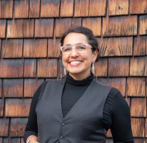 A person with glasses smiles in front of a wooden shingle wall. They have dark hair and are wearing a black turtleneck and a gray vest. Sporting large, dangling earrings and a bracelet, they exude confidence reminiscent of someone who just aced their Berkeley Journalism class.