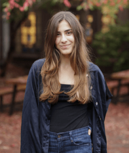 Anabel stands outdoors, her long brown hair flowing, wearing a dark blue jacket over a black top and blue jeans. The background features a blurred scene with wooden benches and autumn leaves on the ground, with green and reddish foliage in the distance.