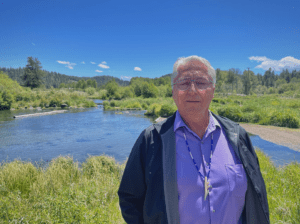 A man with gray hair and glasses, wearing a purple shirt, a beaded necklace, and a dark jacket, stands in front of a scenic grassy riverside with a clear blue sky and forested hills in the background—capturing the essence of Berkeley Journalism.