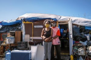 A couple stands in front of a makeshift shelter. The woman wears a yellow mask and the man, a grey mask. The shelter, reminiscent of scenes often covered by Berkeley Journalism, is constructed from various materials including tarps and wooden panels, and it appears cluttered with household items. The sky is clear and blue.