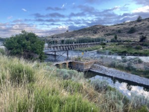 A scenic view of a dam with a bridge structure spanning a river, surrounded by grassy hills and vegetation. The sky above is partly cloudy with hints of blue, and there is a calm section of water in the foreground, setting the perfect scene for any Berkeley Journalism piece on nature's tranquility.